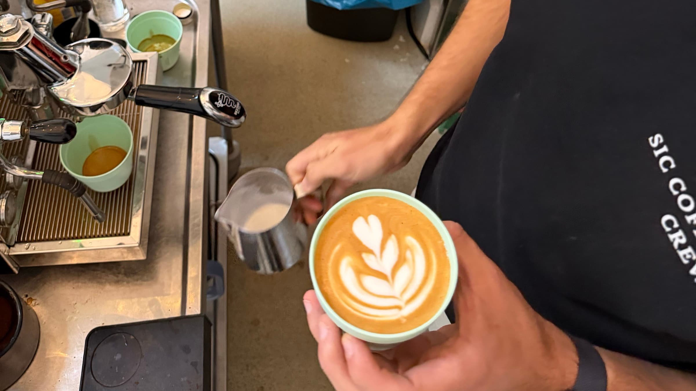 Barista gießt eine Latte-Art-Rosette in einen Becher, im Hintergrund die Siebträgermaschine