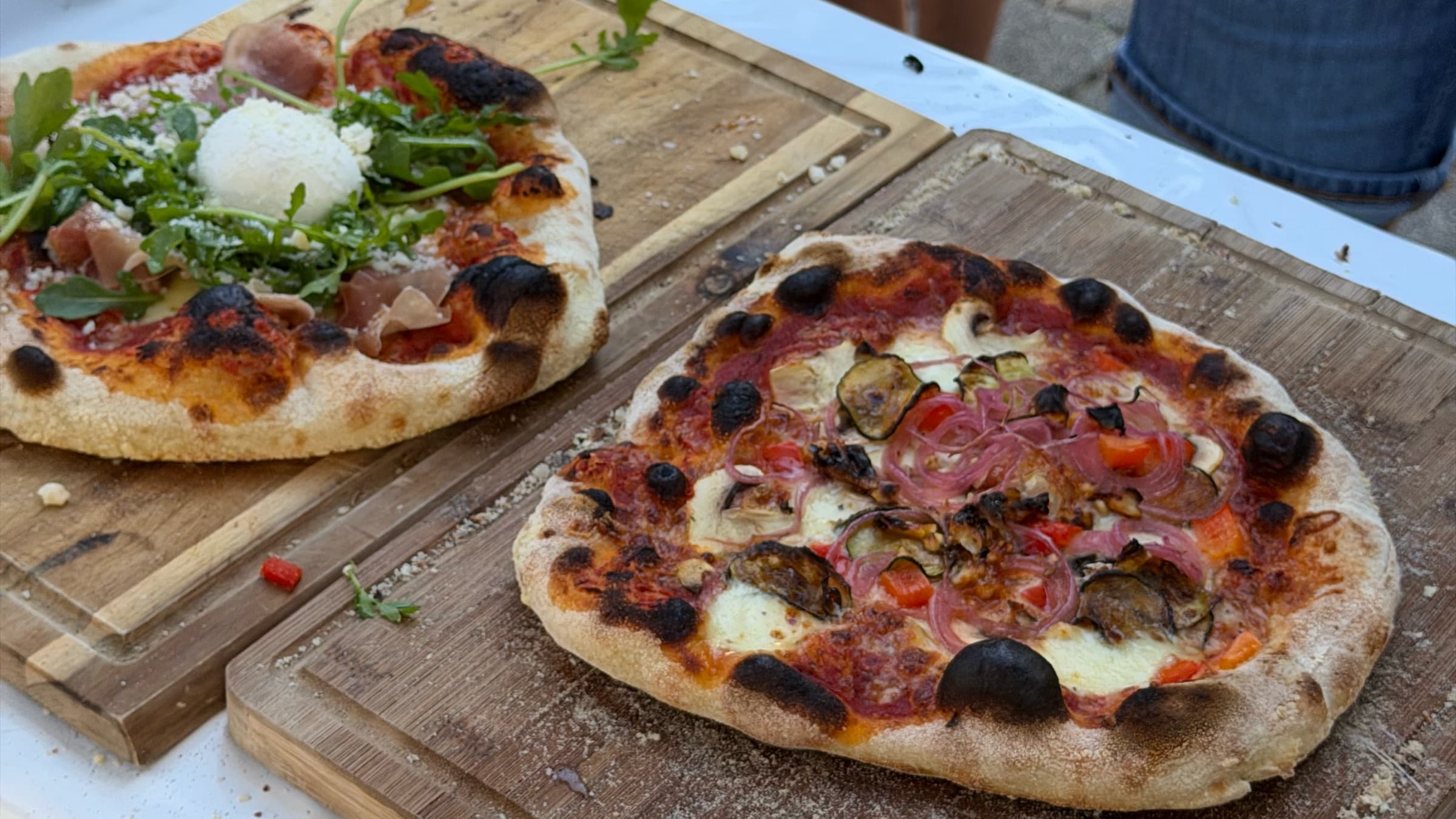 Zwei frisch gebackene Pizzen auf Holzbrettern, dahinter warten die Gäste