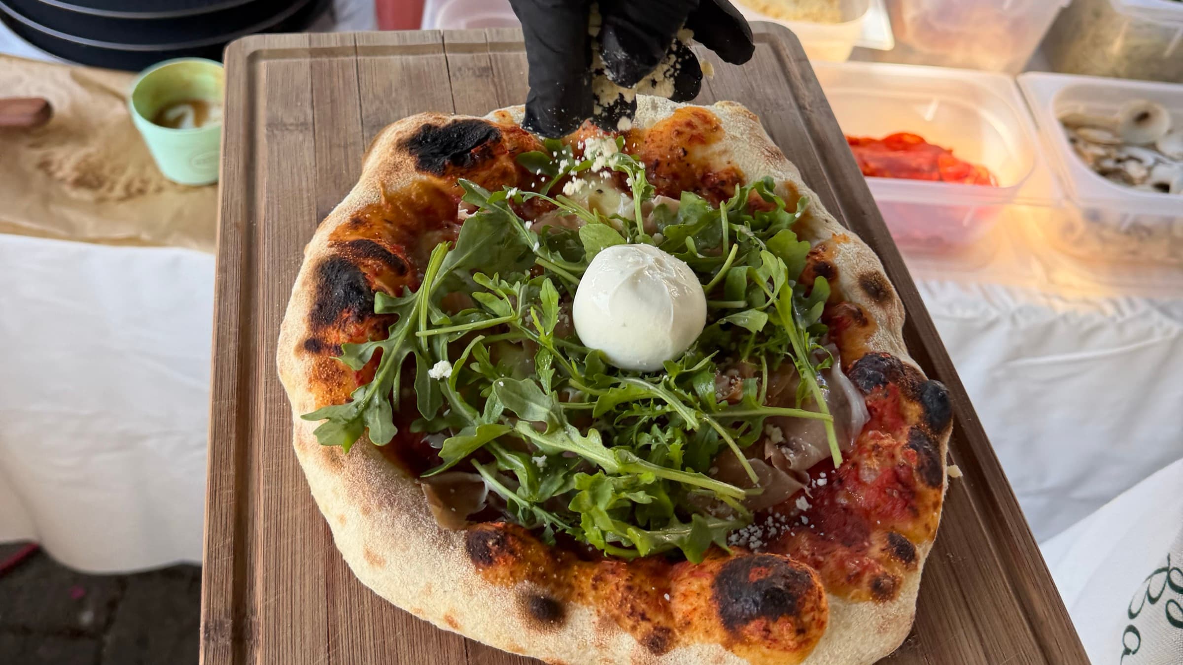 Pizza mit Rucola und Burrata wird frisch belegt, im Hintergrund die Zutatenstation